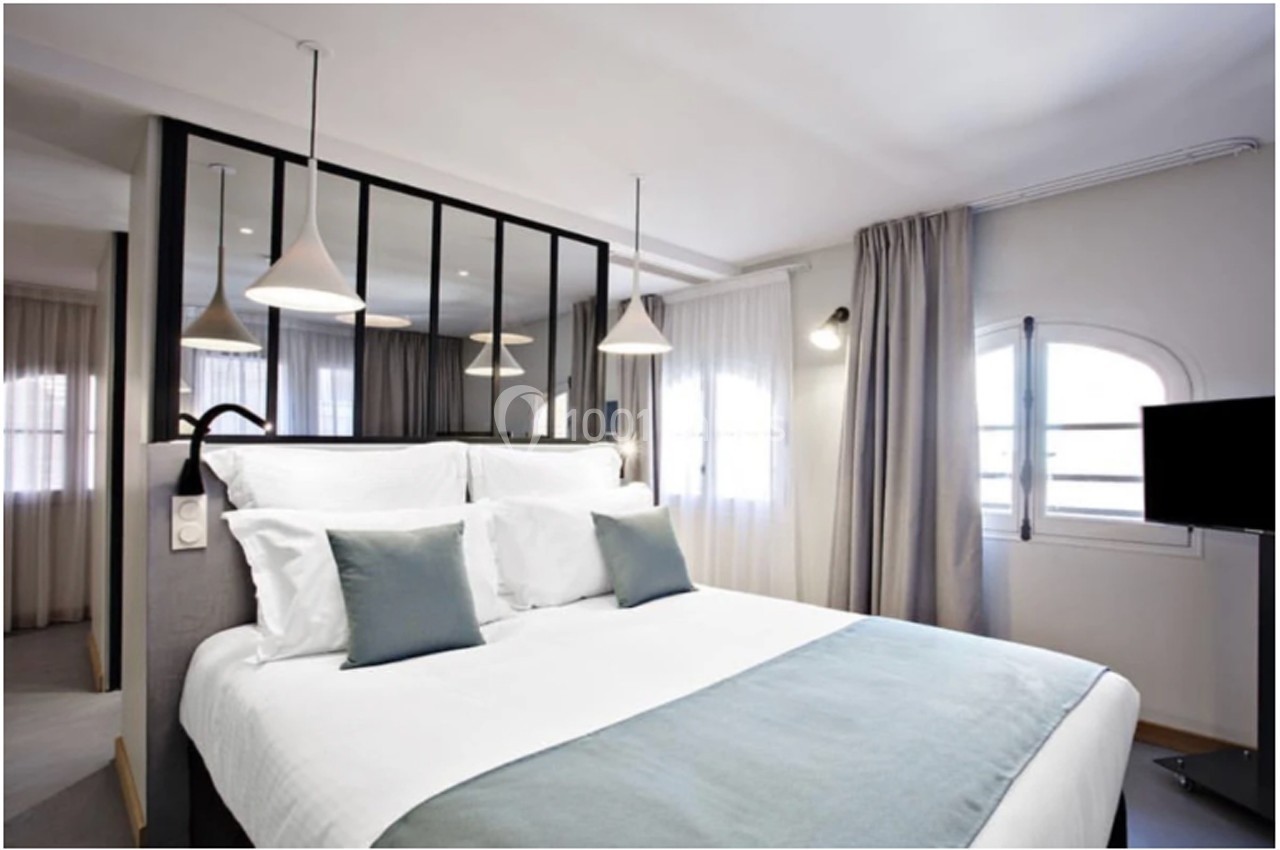 Chambre lumineuse avec lit double, coussins bleus, suspension moderne et fenêtres avec rideaux clairs.