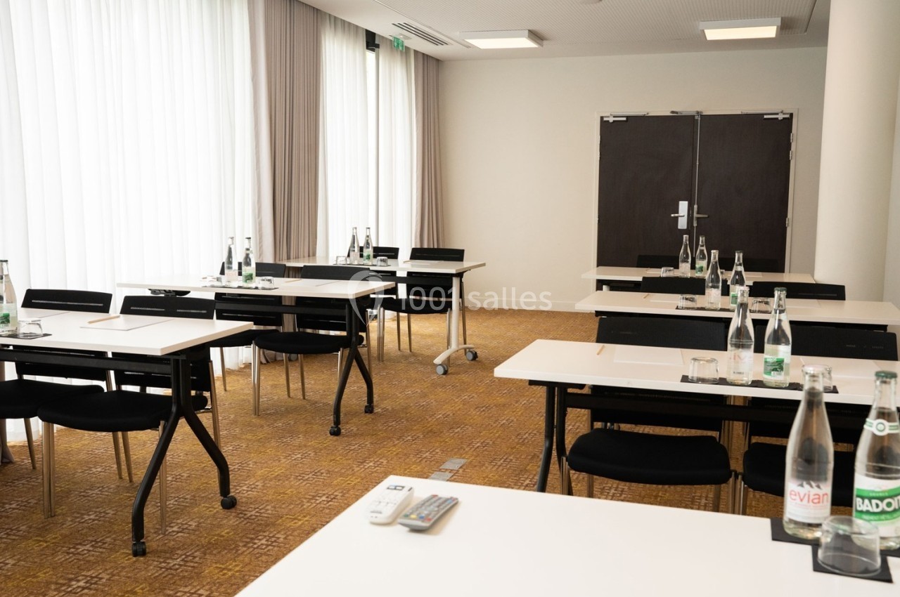 Salle de réunion lumineuse avec des tables alignées, chaises noires et bouteilles d'eau disposées sur les tables.