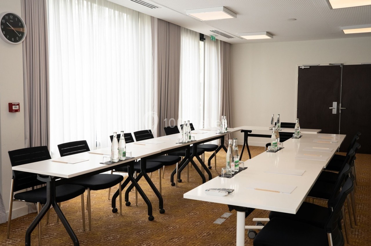 Salle de réunion lumineuse avec deux tables blanches alignées, chaises noires et bouteilles d'eau disposées.