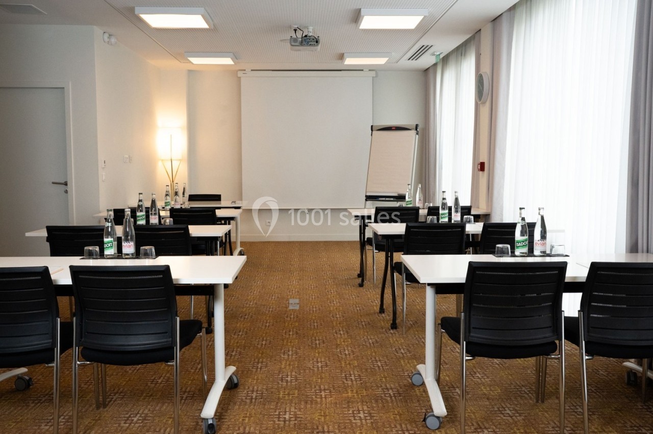 Salle de réunion lumineuse avec des tables, chaises, un tableau blanc et un écran de projection.