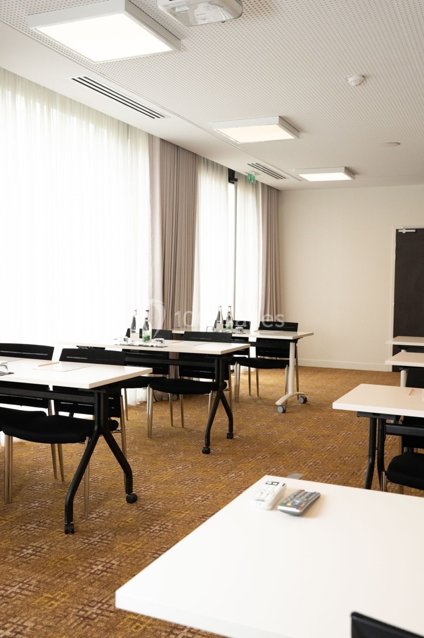 Salle de réunion lumineuse avec tables blanches, chaises noires, bouteilles d'eau et rideaux beige clair.