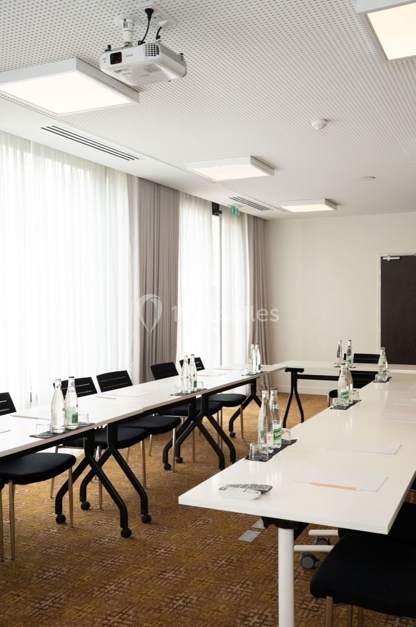 Salle de réunion lumineuse avec tables blanches disposées en U, chaises noires et bouteilles d'eau sur les tables.