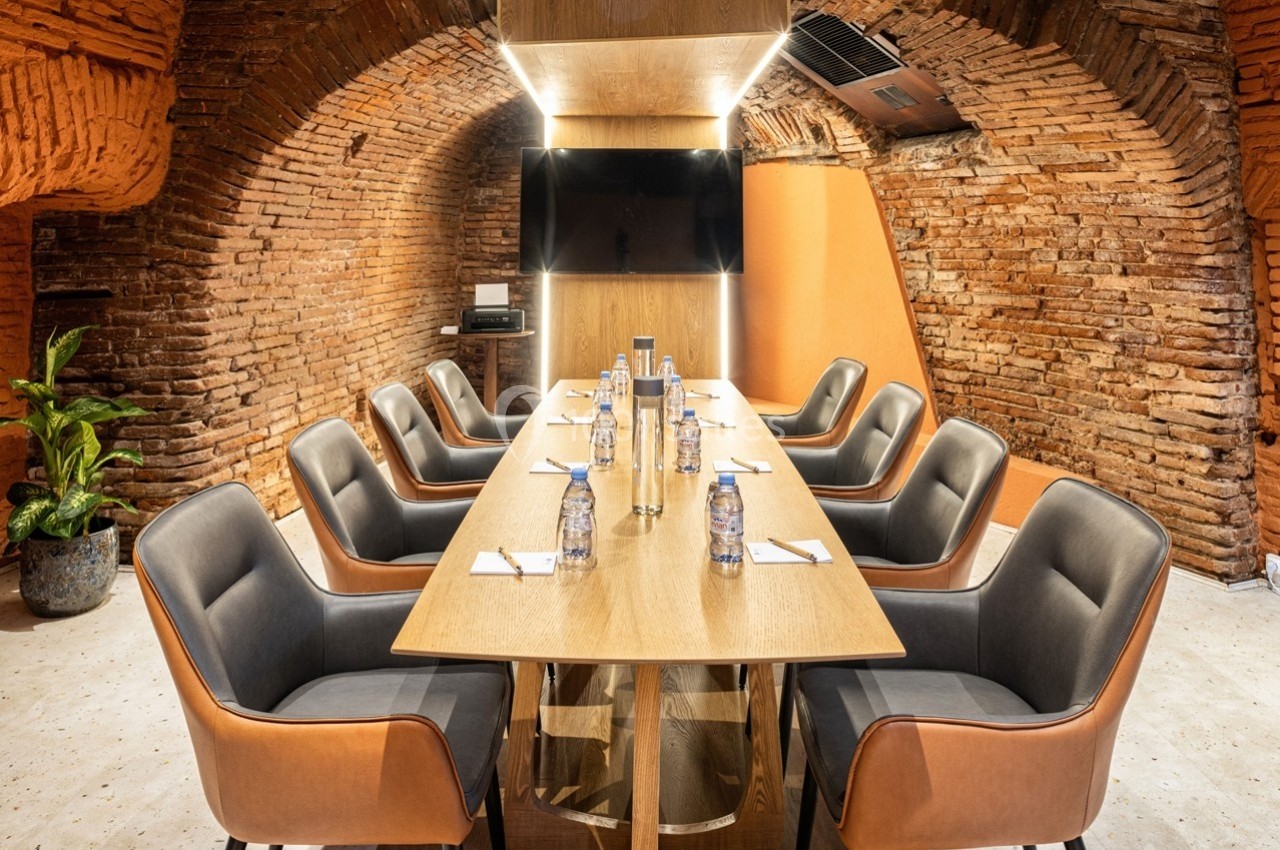 Salle de réunion voûtée en briques avec table en bois, chaises modernes, écran mural et bouteilles d'eau alignées.