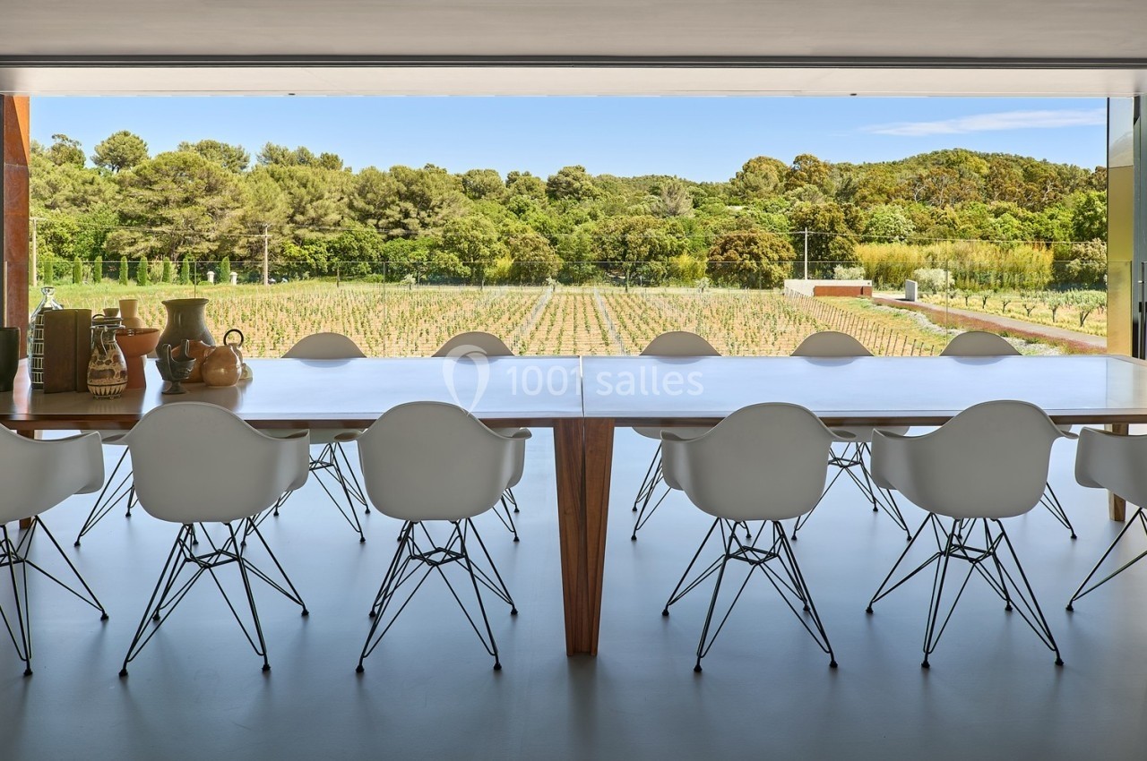Salle à manger moderne avec une grande table en bois, des chaises blanches et vue sur un vignoble et des arbres.
