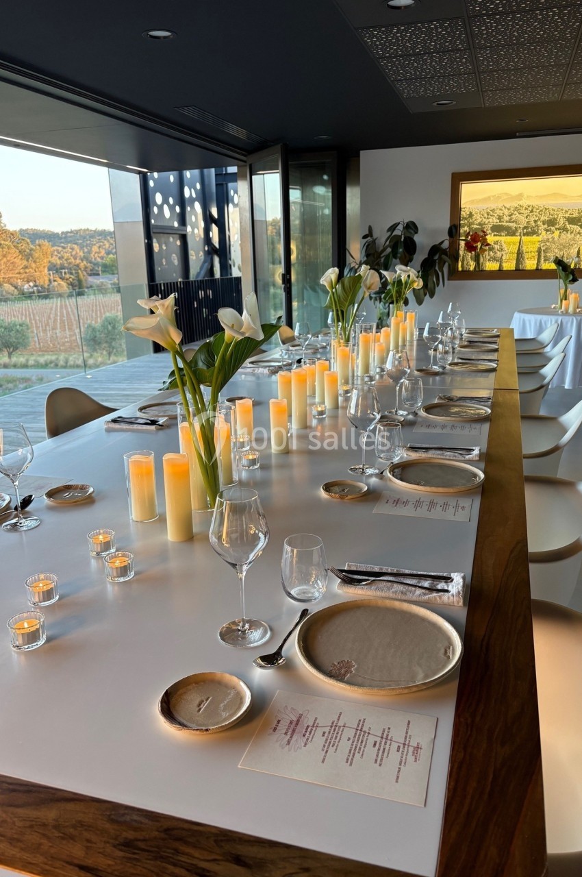 Grande table élégamment dressée avec bougies, fleurs blanches et vue sur un paysage de vignobles à travers une baie vitrée.