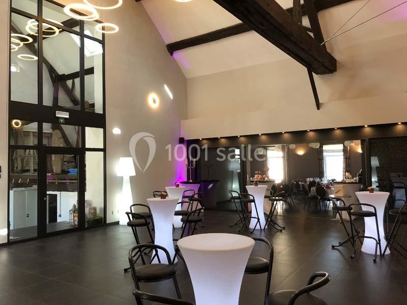 Salle moderne avec poutres apparentes, tables hautes éclairées et grandes baies vitrées laissant entrer la lumière naturelle.