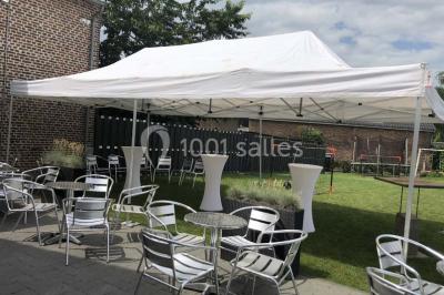 Tente blanche avec toit pointu et bordure rouge installée dans un jardin arboré, entourée de tables et chaises.