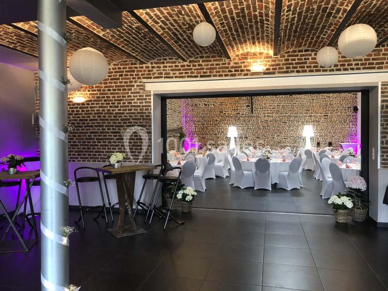 Salle de réception en briques apparentes avec tables dressées, chaises blanches et décorations lumineuses.