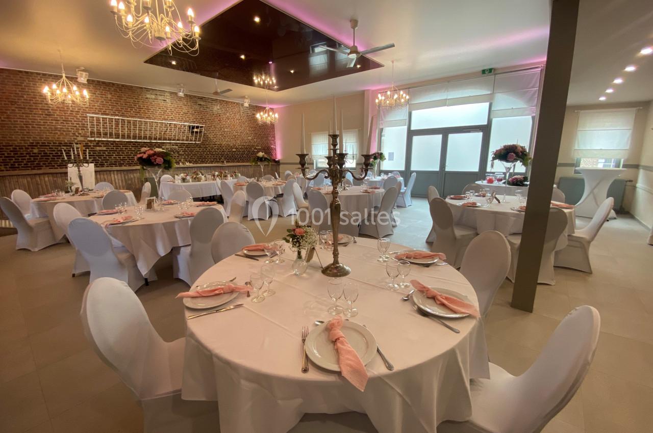 Salle de réception décorée avec des tables rondes dressées, nappes blanches et chaises houssées, éclairage chaleureux.