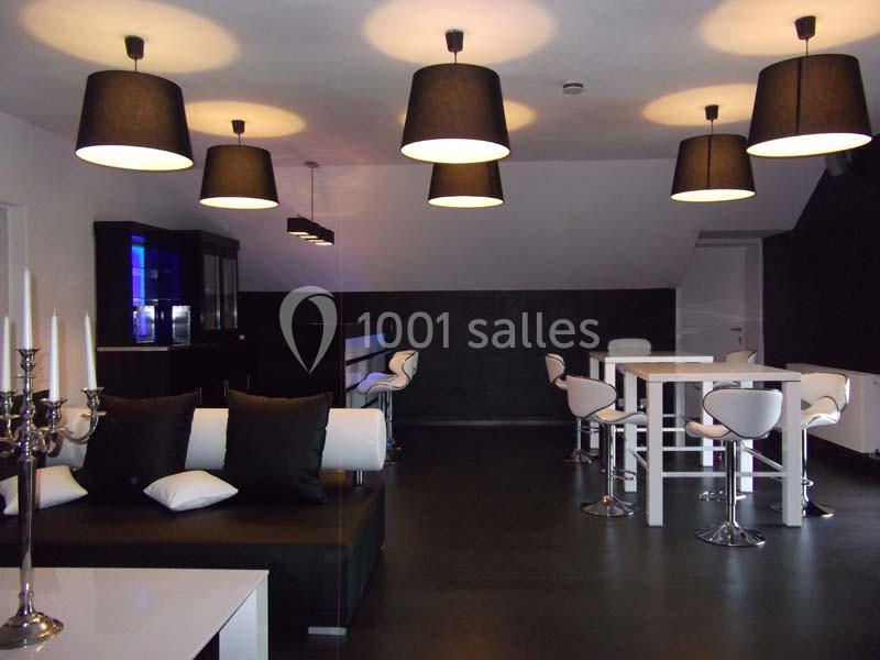 Salle moderne avec éclairage suspendu, mobilier noir et blanc, et un bar avec tabourets hauts en arrière-plan.