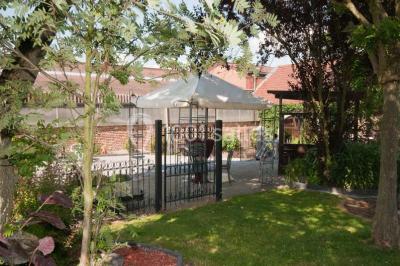 Tente blanche avec toit pointu et bordure rouge installée dans un jardin arboré, entourée de tables et chaises.