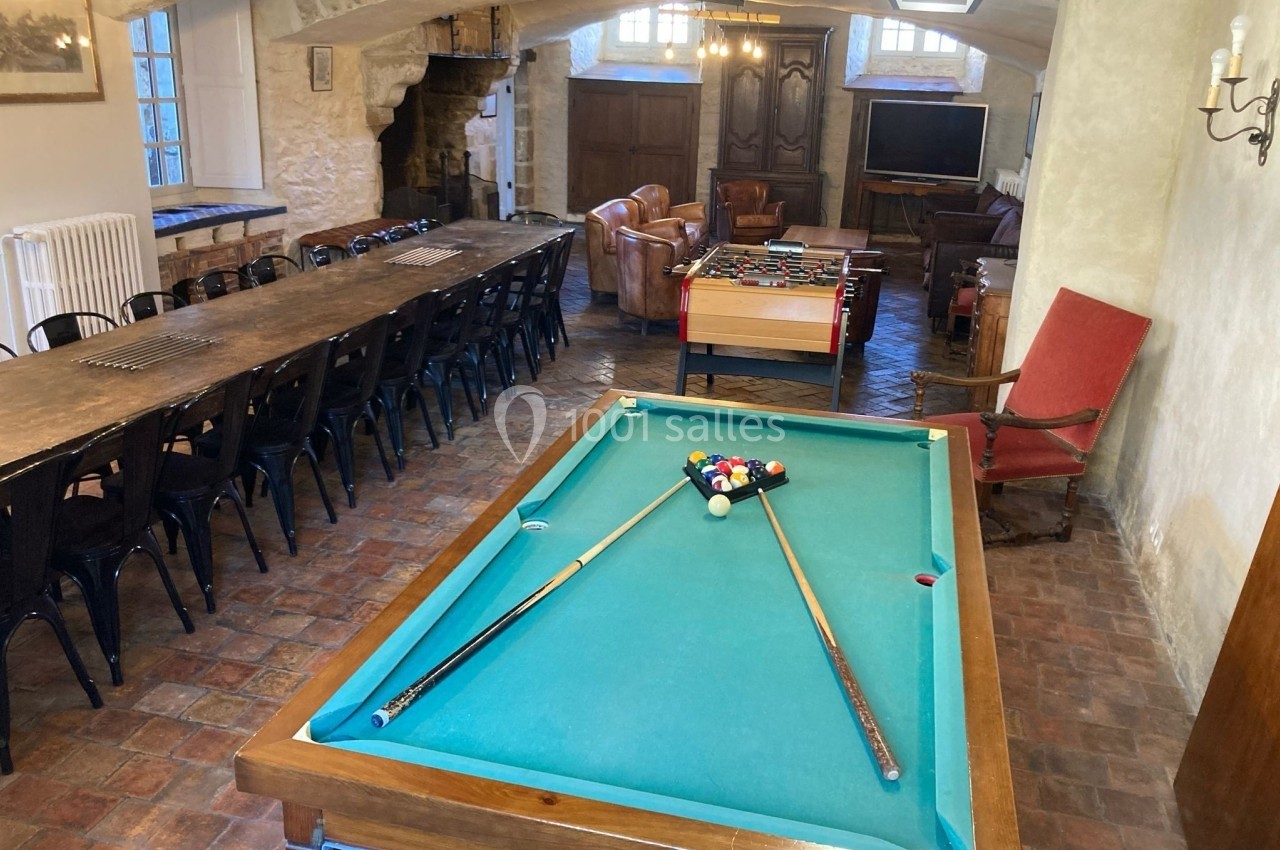 Salle avec une table de billard, baby-foot, longue table en bois, fauteuils et murs en pierre apparente.