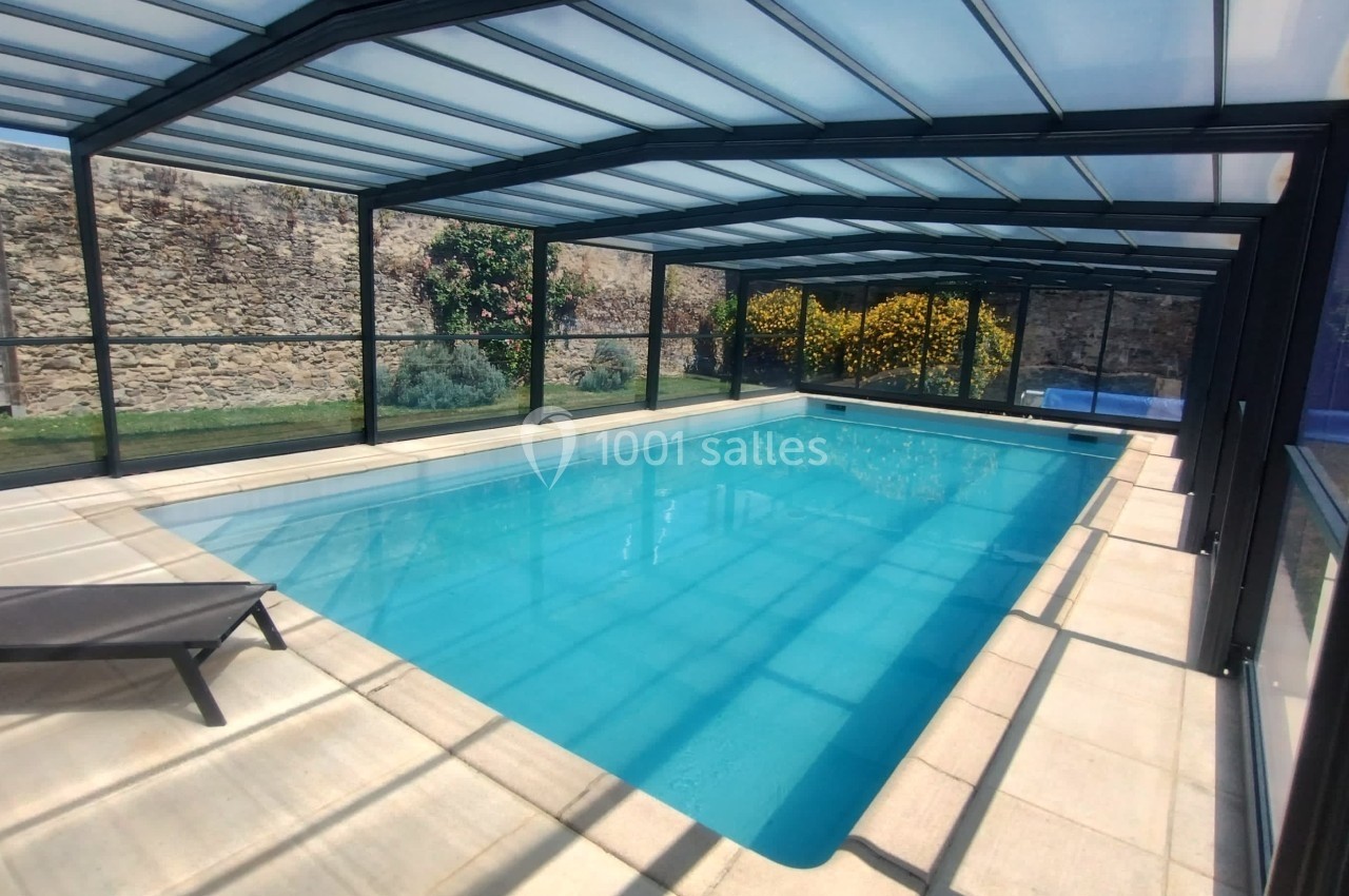 Piscine couverte avec structure vitrée, entourée de murs en pierre et d'un jardin fleuri en arrière-plan.