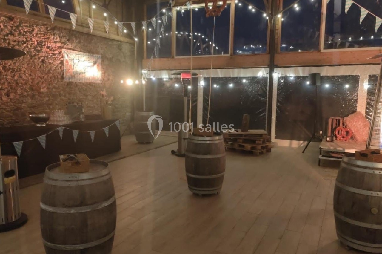 Salle décorée pour un événement, avec tonneaux en guise de tables, guirlandes lumineuses et murs en pierre apparente.