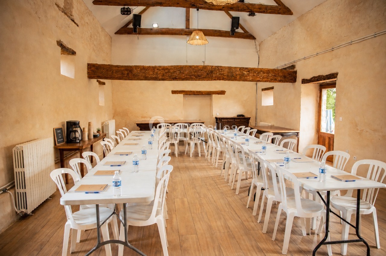 Salle de réunion lumineuse avec tables alignées, chaises blanches en plastique et poutres apparentes au plafond.