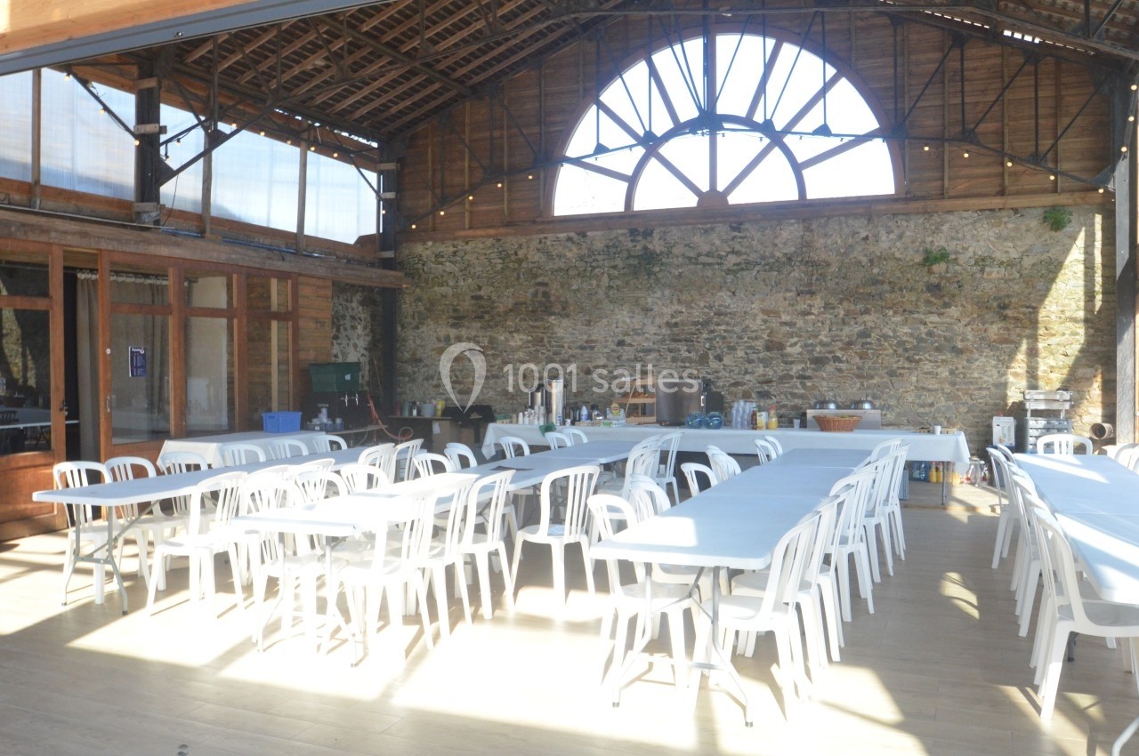 Salle lumineuse avec de grandes baies vitrées, murs en pierre et tables blanches alignées avec des chaises.