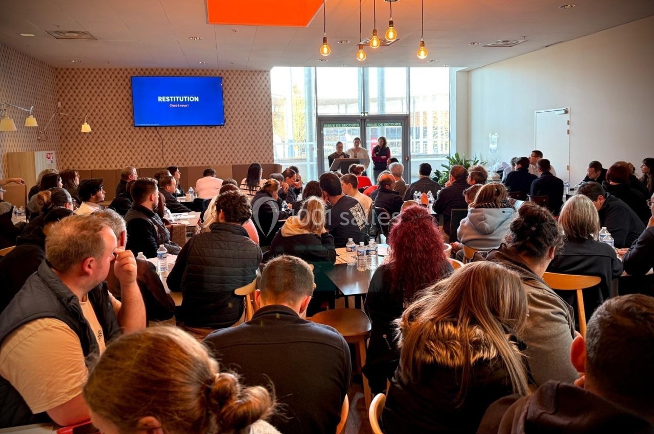 Salle comble avec des participants assis écoutant une présentation sur scène devant un écran affichant ’Restitution’.
