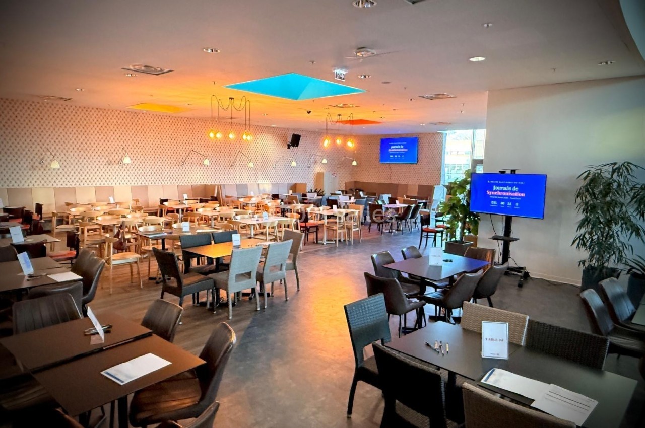 Salle de restaurant spacieuse avec tables et chaises disposées, éclairage chaleureux et écran de présentation allumé.