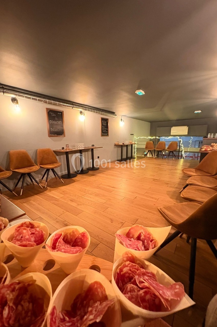Salle lumineuse avec parquet, chaises alignées, tables hautes et cônes de charcuterie en premier plan.