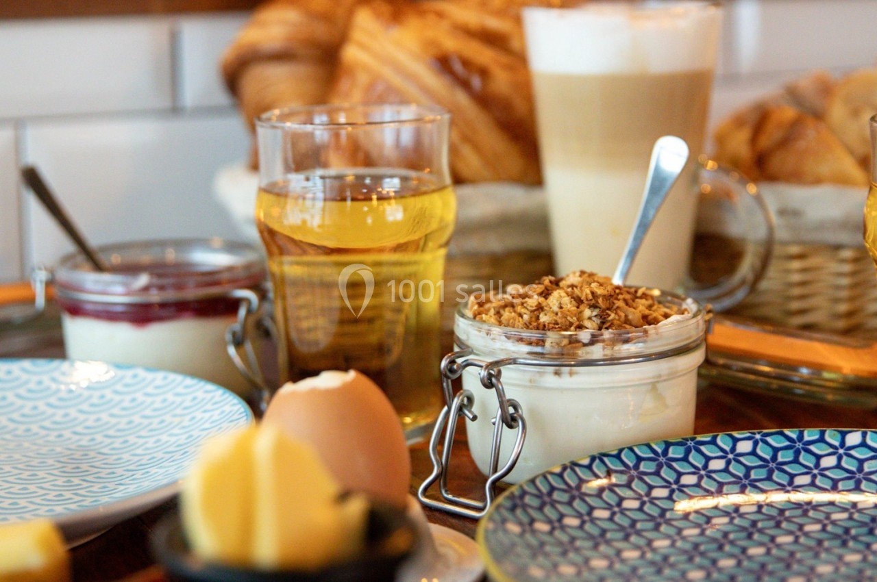 Petit-déjeuner composé de yaourt au granola, jus, café, viennoiseries, beurre et œuf à la coque sur une table.