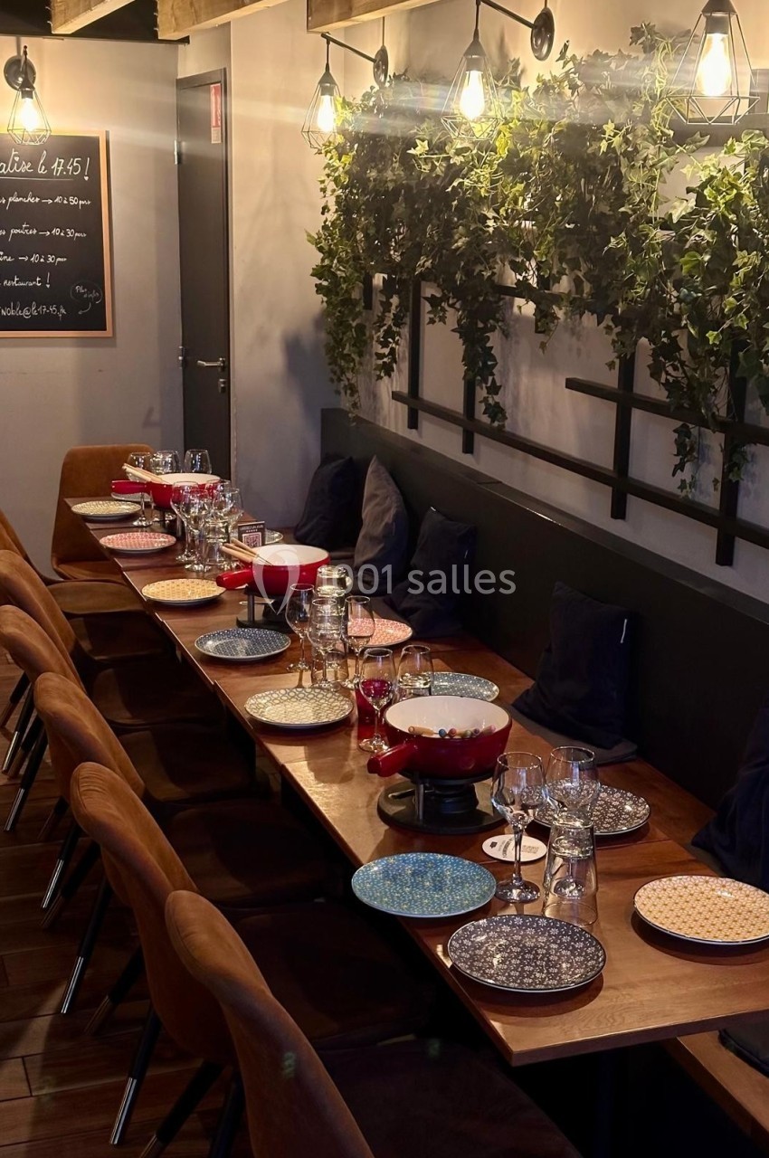 Table en bois dressée pour un repas, avec assiettes colorées, verres et fondue, dans une salle décorée de plantes murales.