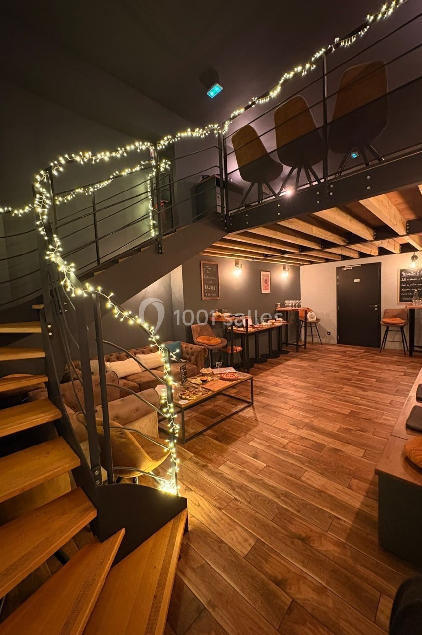 Escalier en bois menant à une mezzanine, avec guirlandes lumineuses et espace salon au décor chaleureux en dessous.