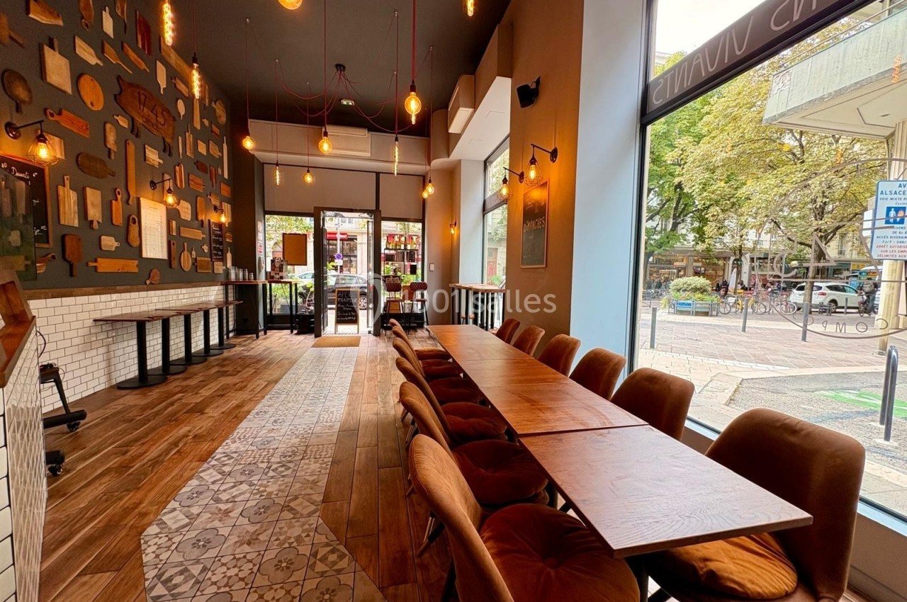 Intérieur d'un café avec une grande table en bois, des chaises rembourrées et une vue sur l'extérieur par une baie vitrée.
