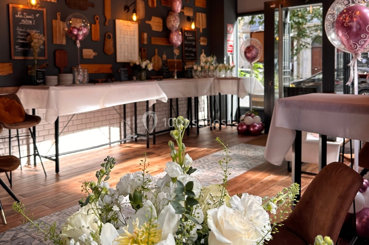 Salle décorée pour un événement avec ballons roses, nappes blanches et bouquet de fleurs blanches au premier plan.