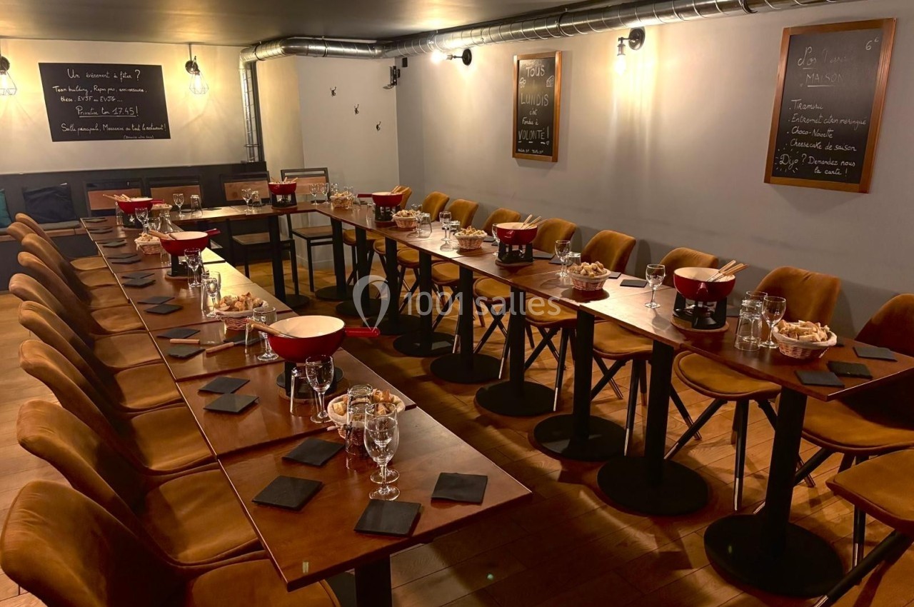 Salle de restaurant avec de longues tables en bois dressées, entourées de chaises en velours et éclairées par des lampes…