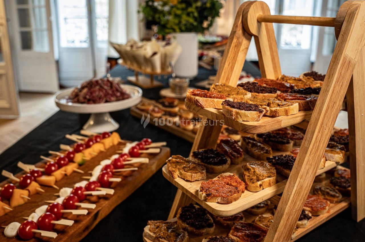 Plateau de toasts variés sur un présentoir en bois, entouré de brochettes apéritives et d'autres mets sur une table.