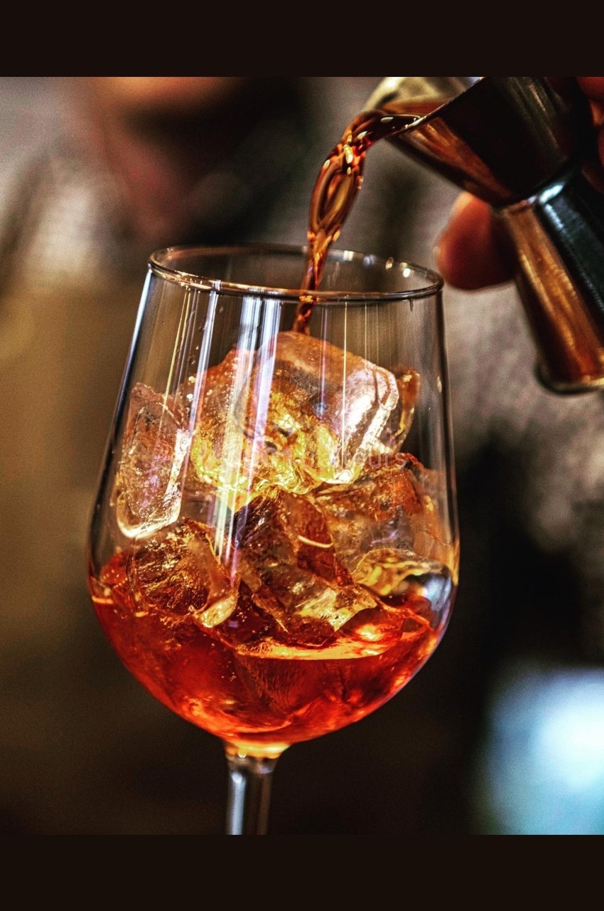Un verre à pied rempli de glaçons, dans lequel une boisson ambrée est versée depuis un doseur métallique.