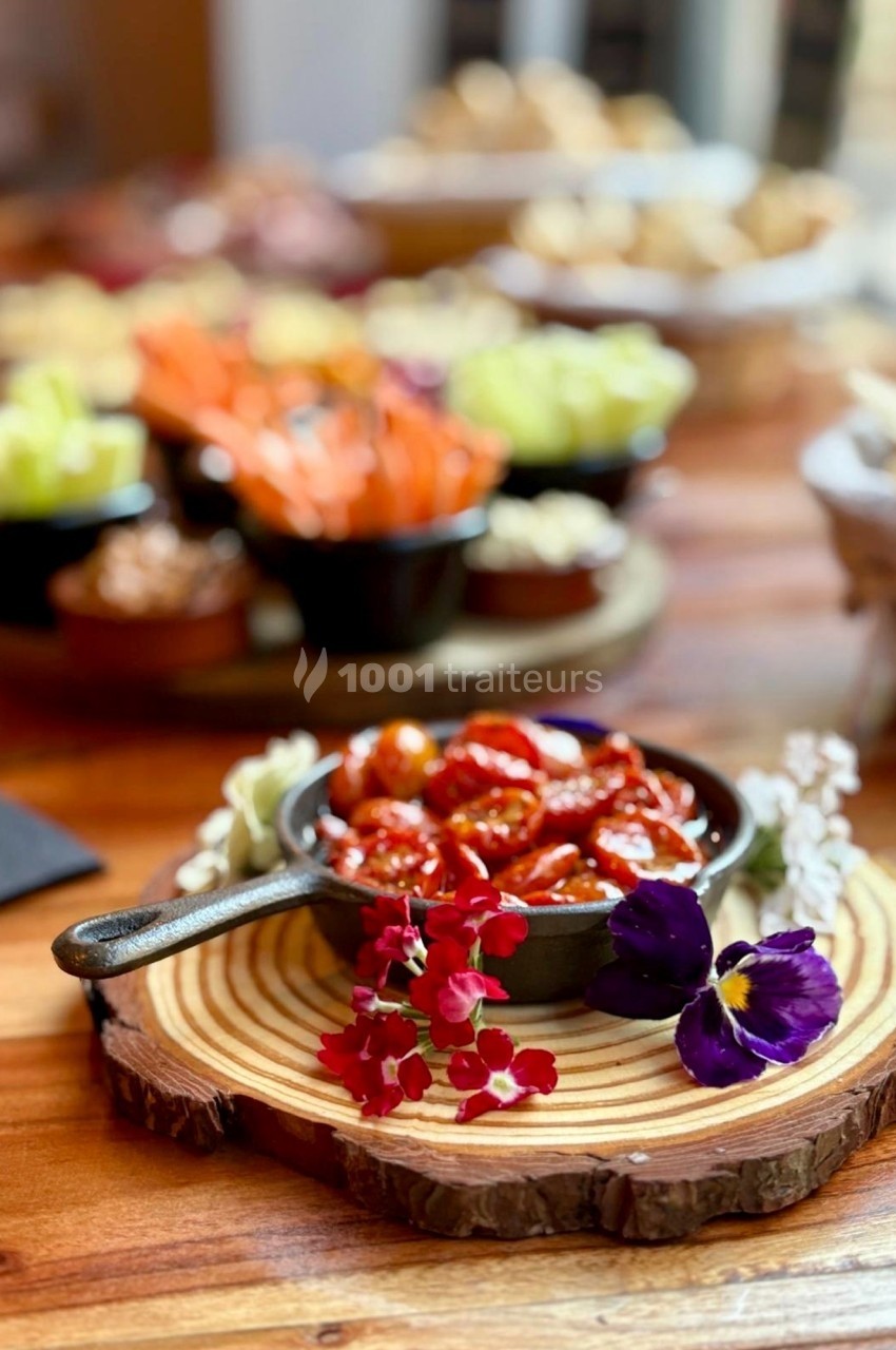 Bol de tomates cerises confites posé sur une planche en bois, entouré de fleurs comestibles, avec des légumes en arrière…