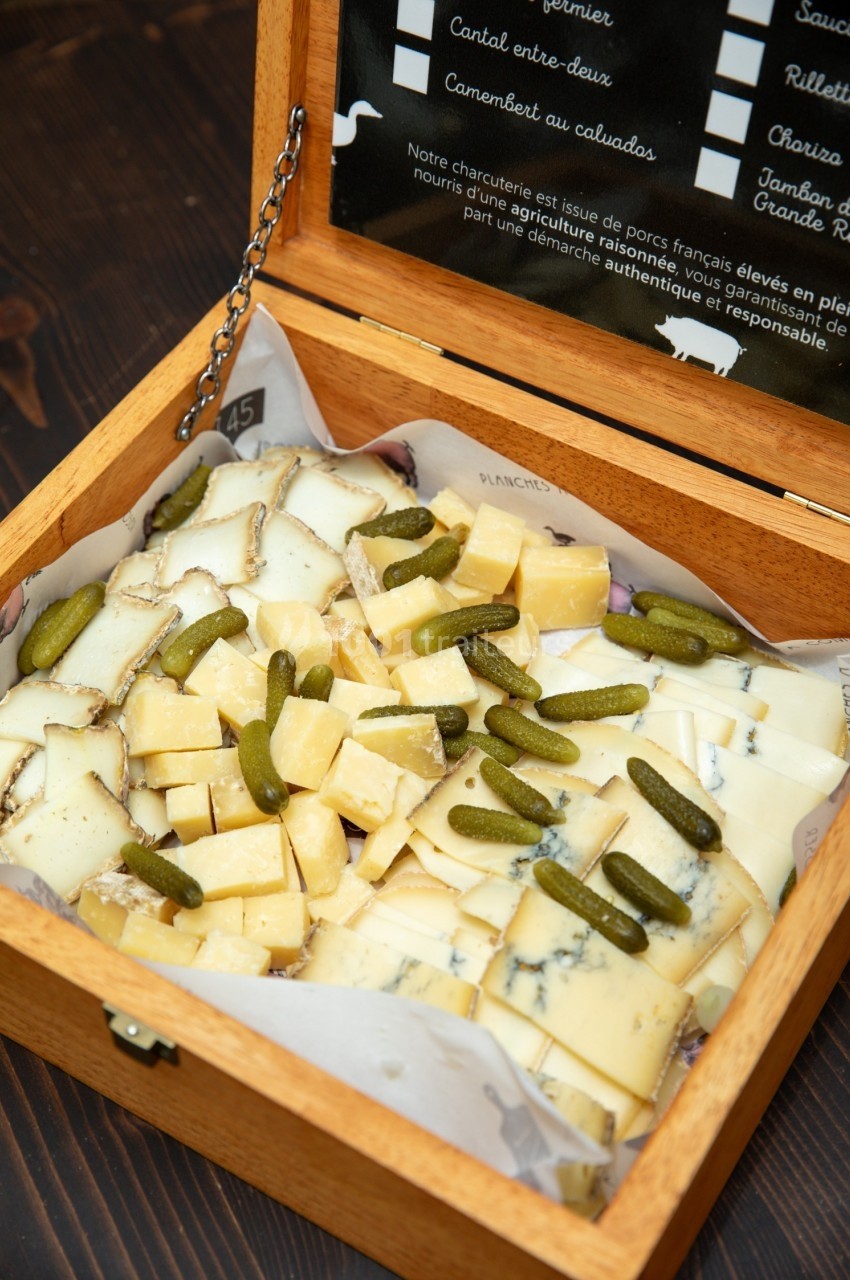 Plateau en bois contenant des cubes de fromage variés et des cornichons, présenté sur une table sombre.