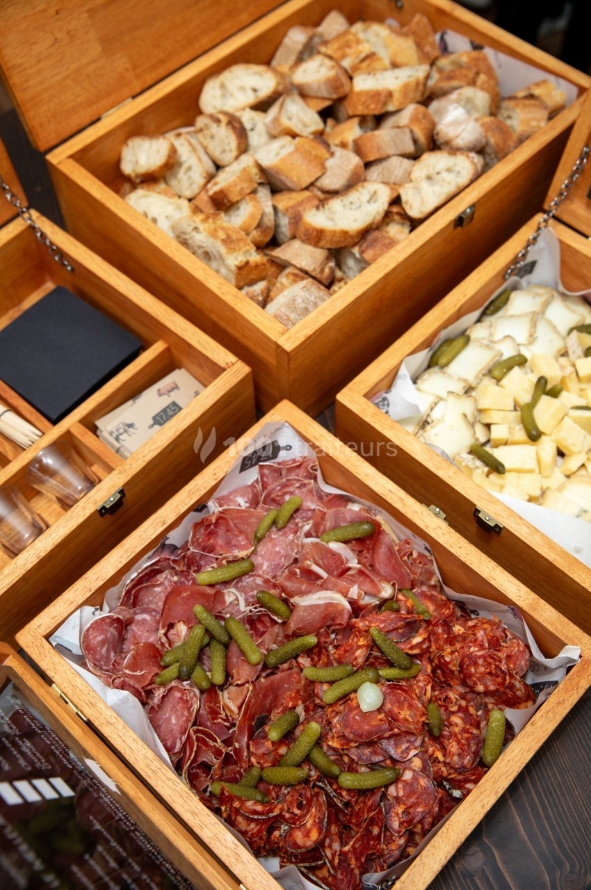 Plateau de charcuterie, fromage, cornichons et pain disposés dans des boîtes en bois ouvertes.