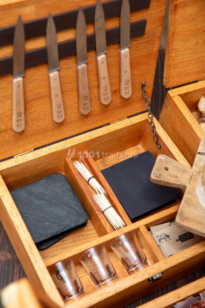 Coffret en bois ouvert contenant des couteaux, des piques en bois, une ardoise, des serviettes et des verres à dégustation.