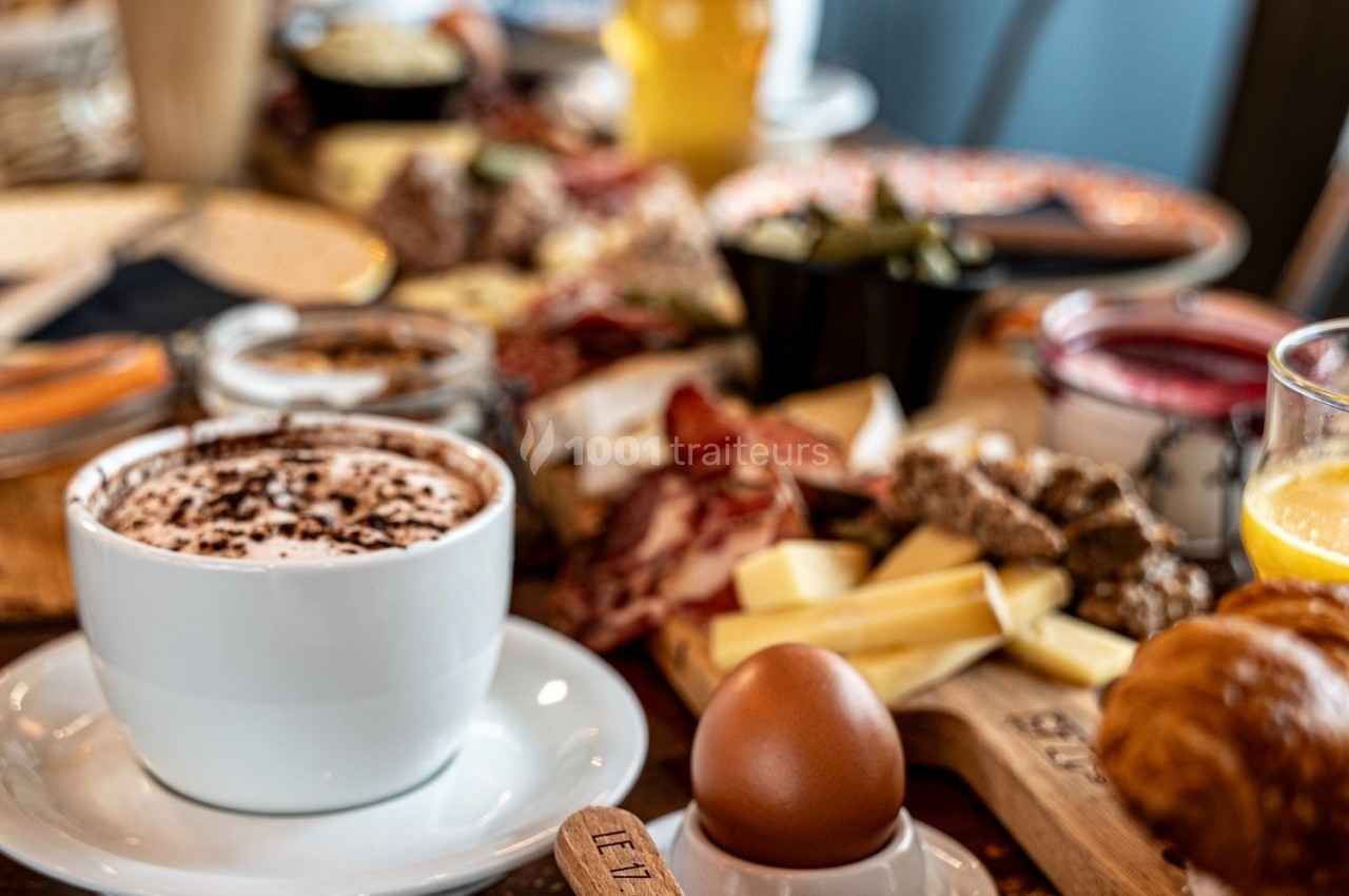 Petit-déjeuner varié avec café, œuf à la coque, viennoiseries, fromages, charcuteries et jus de fruits sur une table.