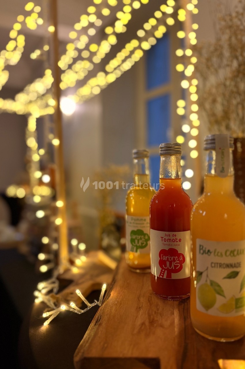 Bouteilles de jus artisanaux disposées sur un présentoir en bois, éclairées par des guirlandes lumineuses.