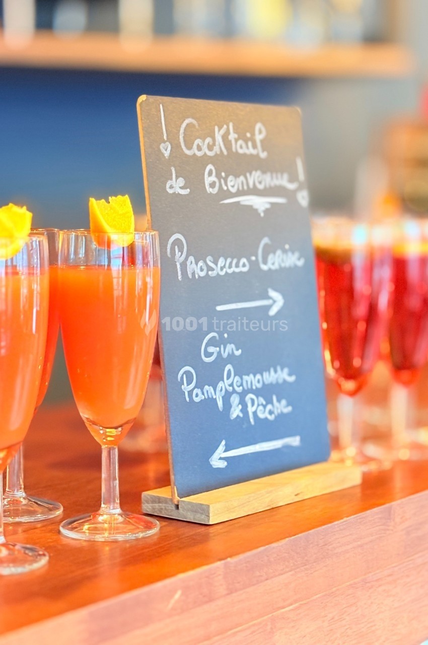 Verres de cocktails colorés alignés sur un comptoir en bois avec une ardoise indiquant les choix disponibles.