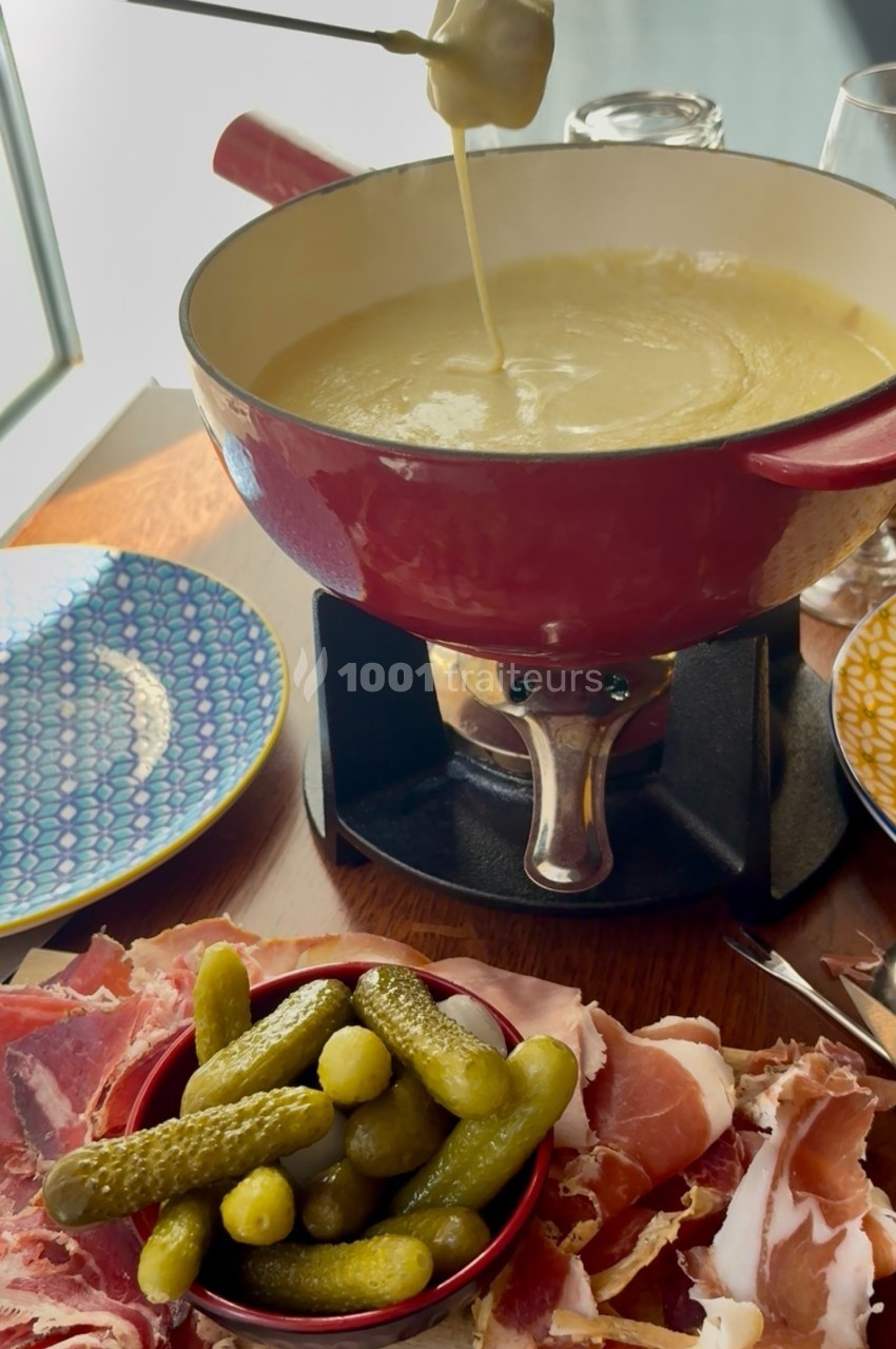 Caquelon de fondue au fromage avec assiette de charcuterie, cornichons et vaisselle colorée sur une table en bois.