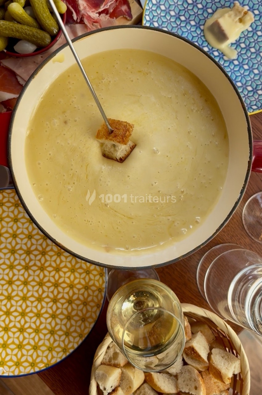 Caquelon de fondue au fromage avec un morceau de pain sur une fourchette, entouré d'accompagnements et de verres.