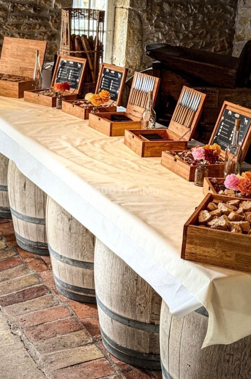 Table dressée sur des tonneaux, avec boîtes en bois contenant couverts, fleurs et aliments dans une salle en pierre.