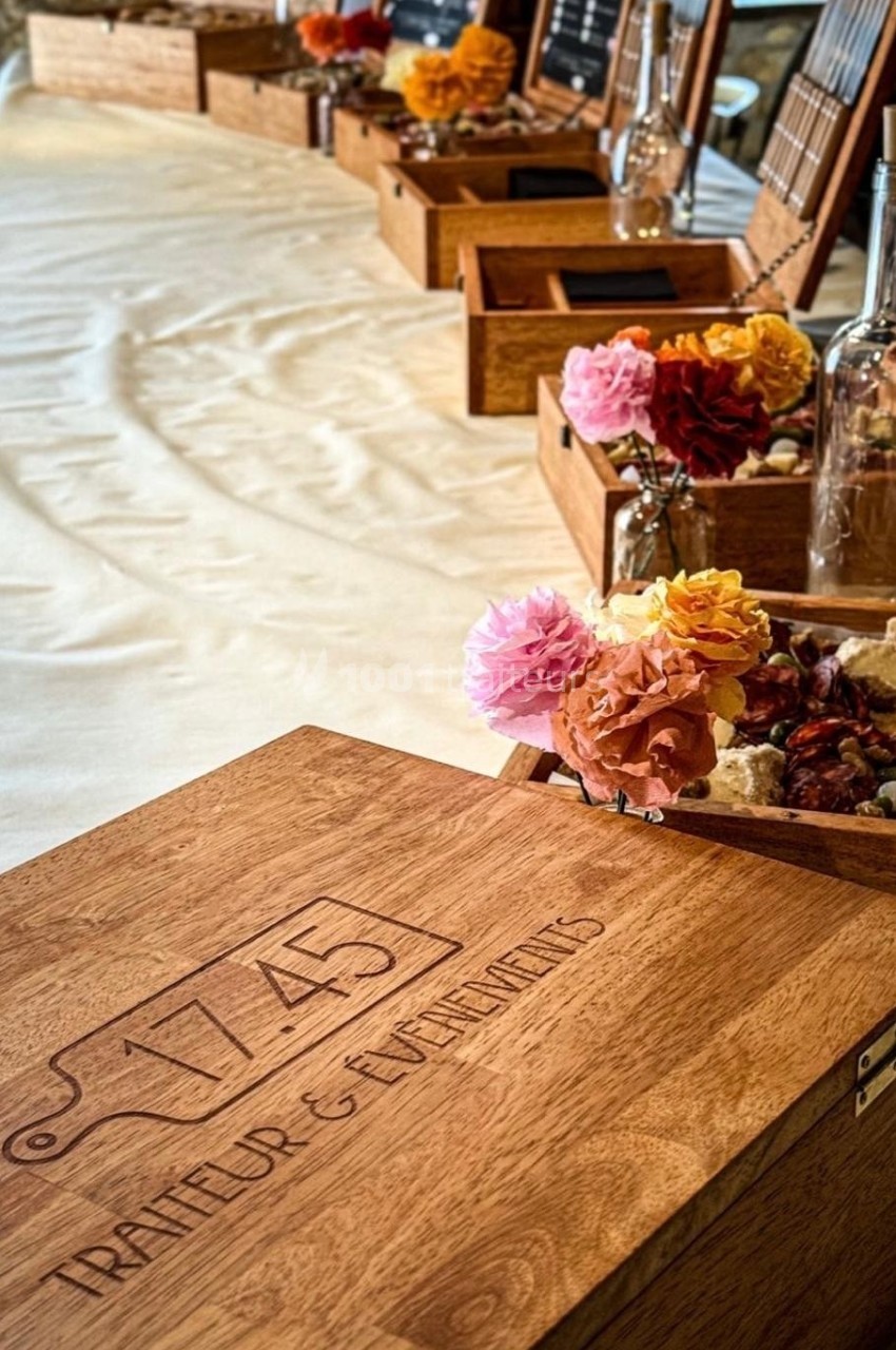 Boîtes en bois gravées ’1745 Traiteur & Événements’ alignées sur une table avec des fleurs et des bouteilles décoratives.