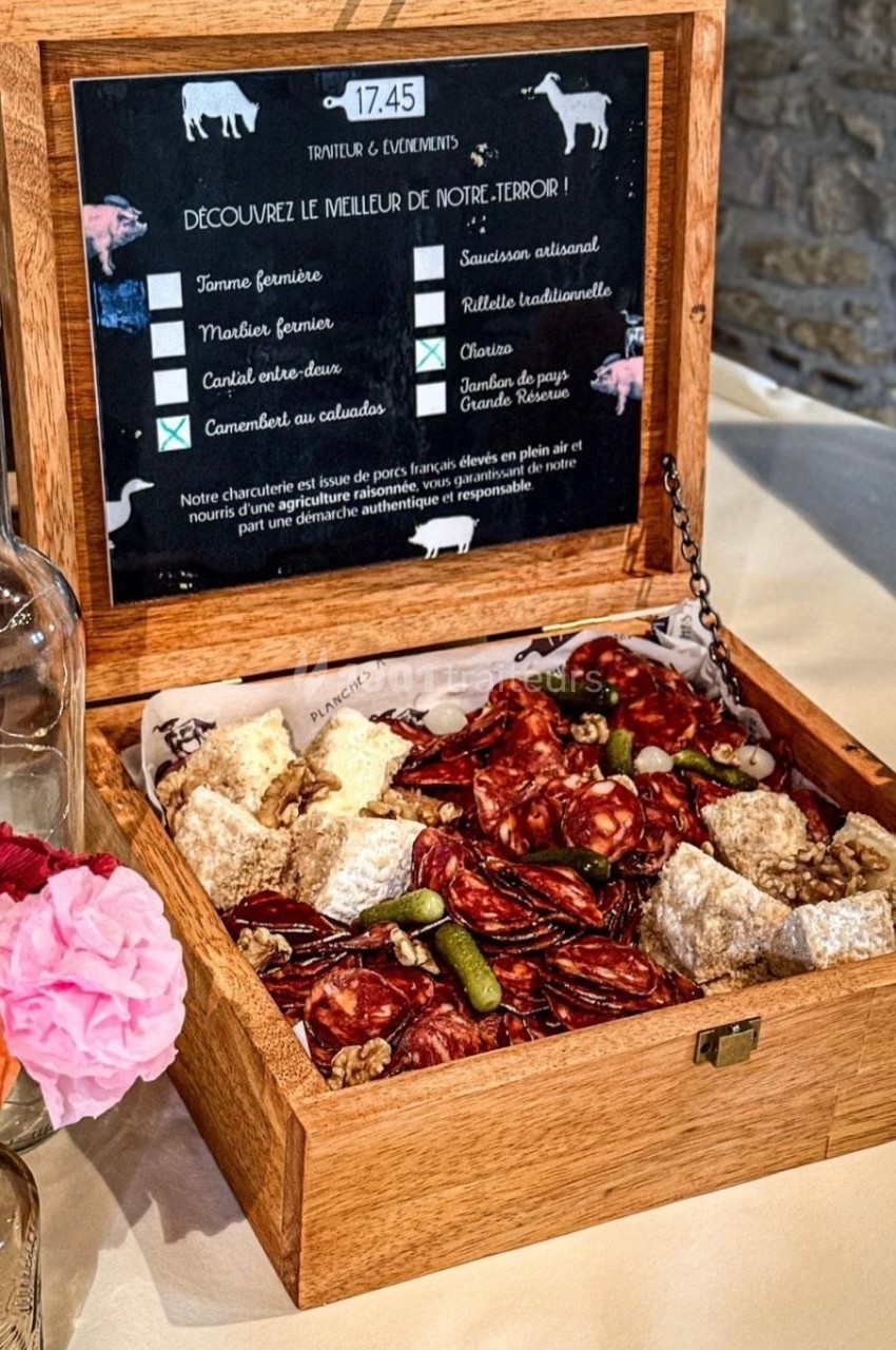 Plateau en bois ouvert contenant des charcuteries variées, cornichons et fromages, posé sur une table avec une fleur rose.