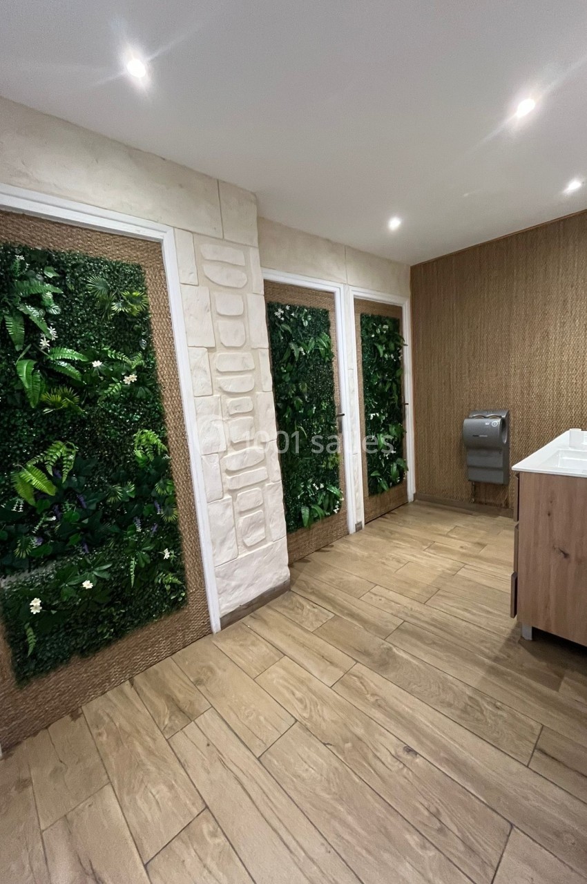 Salle de bain moderne avec murs végétalisés, sol en bois clair, lavabo en bois et distributeur de papier gris.
