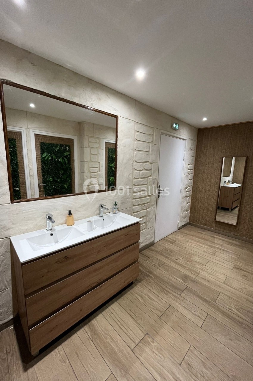 Salle de bain moderne avec double vasque en bois, grand miroir, sol en parquet et murs texturés clairs.