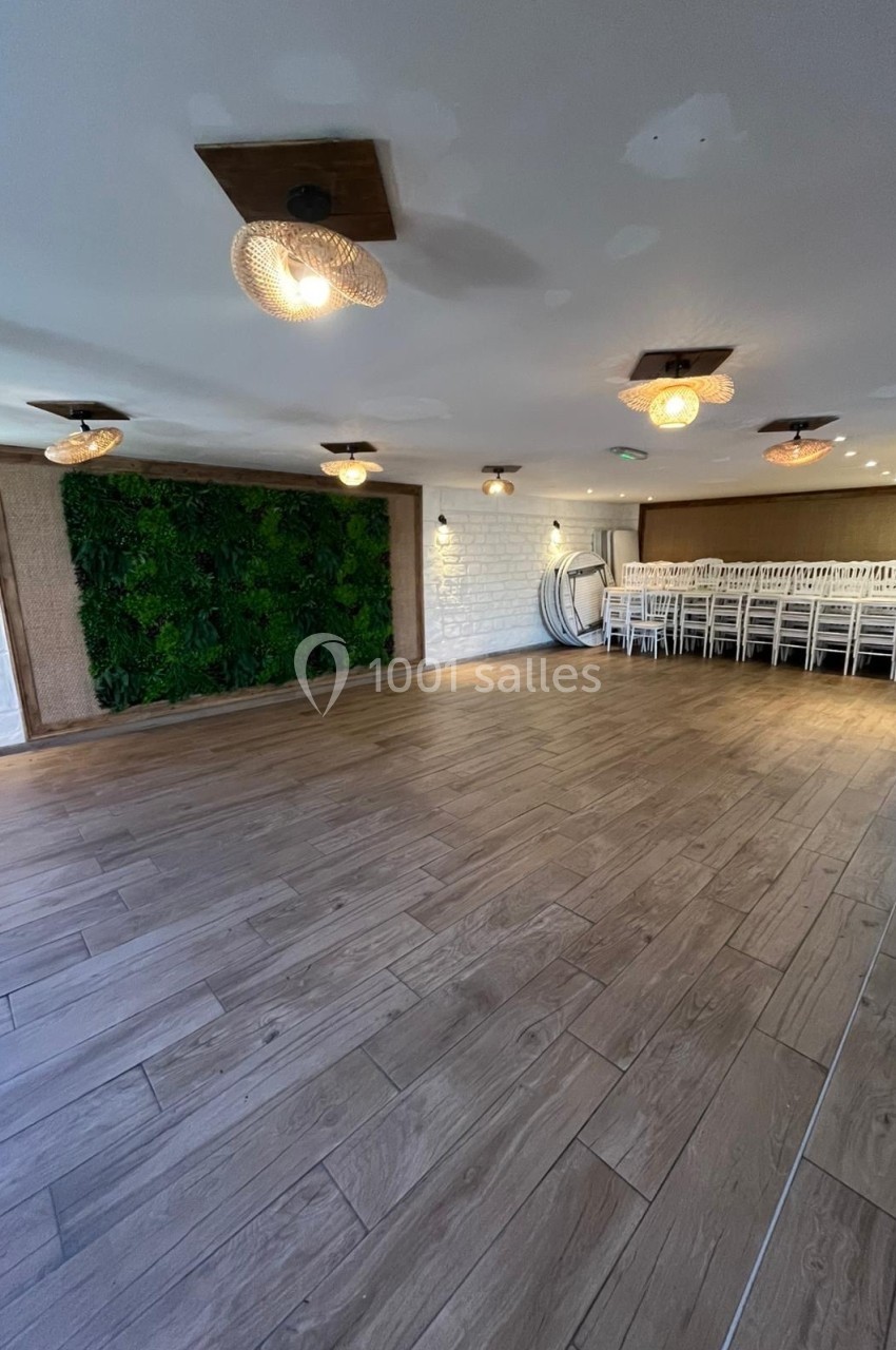 Salle lumineuse avec parquet en bois, mur végétal, chaises empilées et suspensions décoratives au plafond.