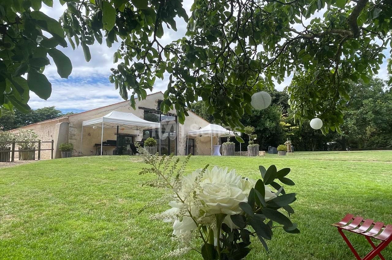 Vue d'un jardin avec pelouse, barnum blanc, bâtiment en pierre et fleurs au premier plan sous un arbre.