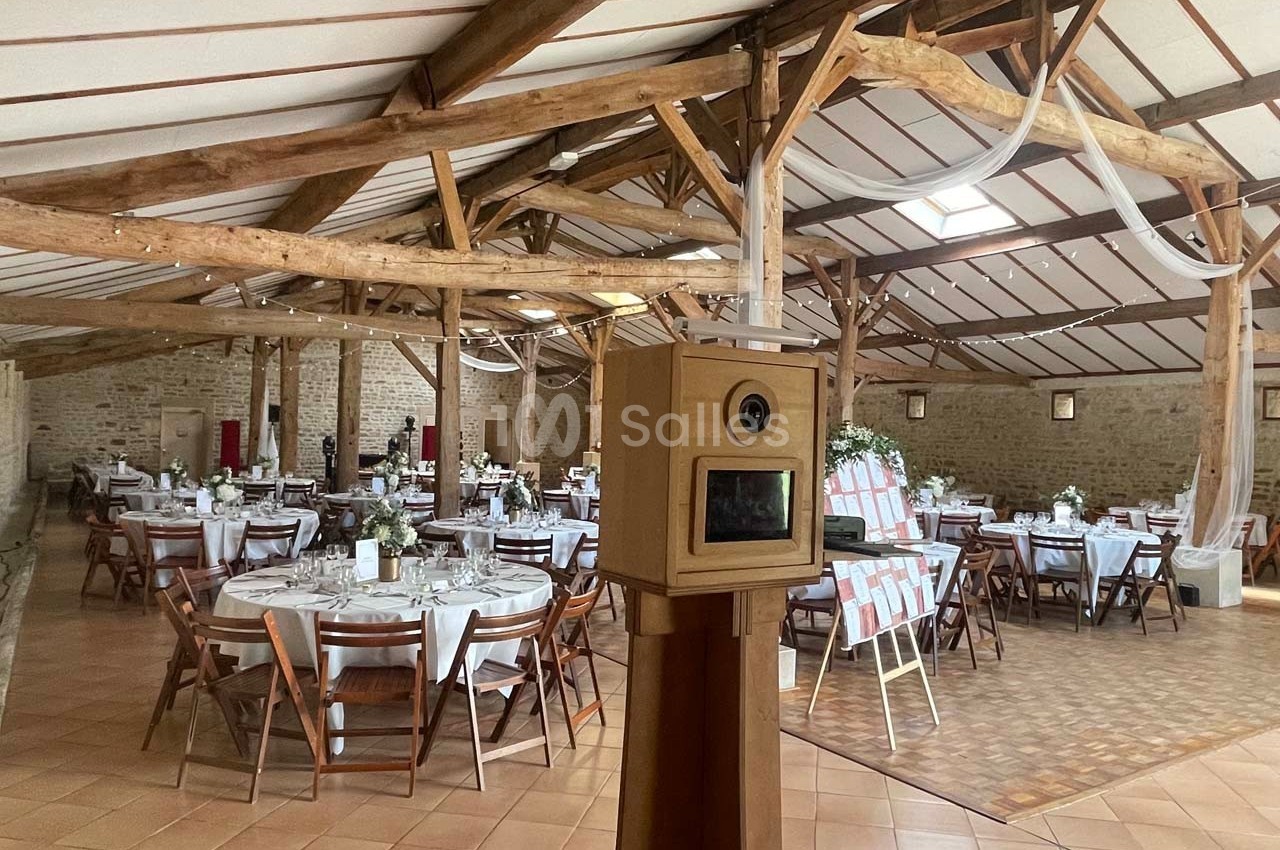 Salle de réception avec tables rondes décorées pour un événement, poutres apparentes et borne photo au premier plan.
