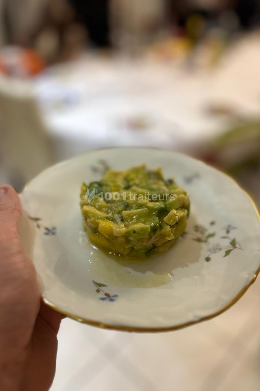 Assiette tenue à la main contenant un tartare d'avocat et de mangue, présentée sur une table floue en arrière-plan.