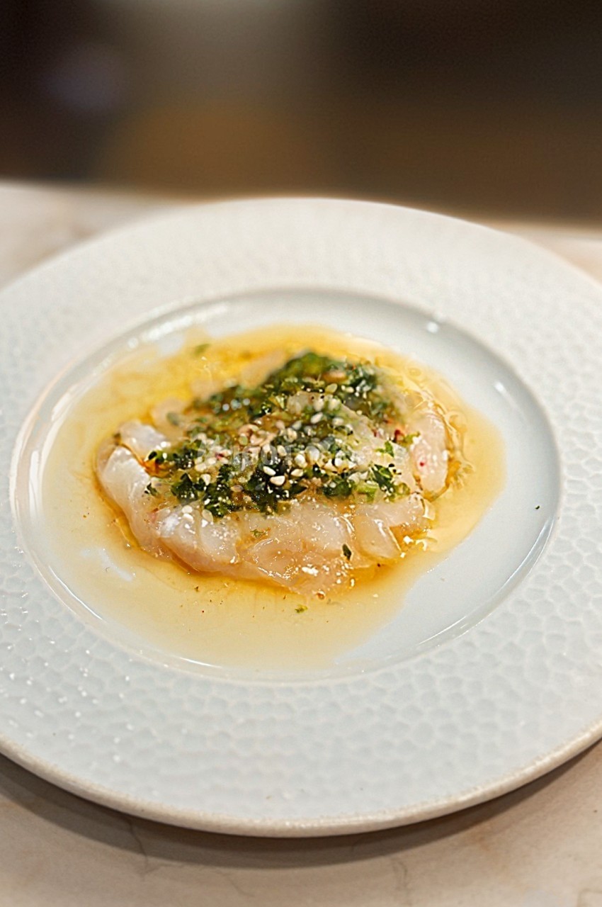 Carpaccio de poisson blanc nappé d'une sauce aux herbes et épices, présenté sur une assiette blanche texturée.